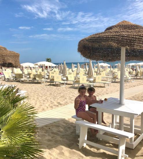 Bambini seduti a un tavolo sulla spiaggia, con ombrelloni e mare sullo sfondo.